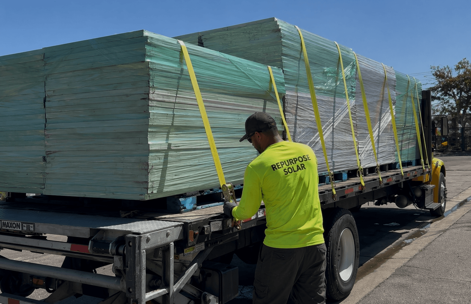 Repurpose Solar crew member securing yellow straps on a flatbed loaded with wrapped, palletized solar panels en route to a recycling facility